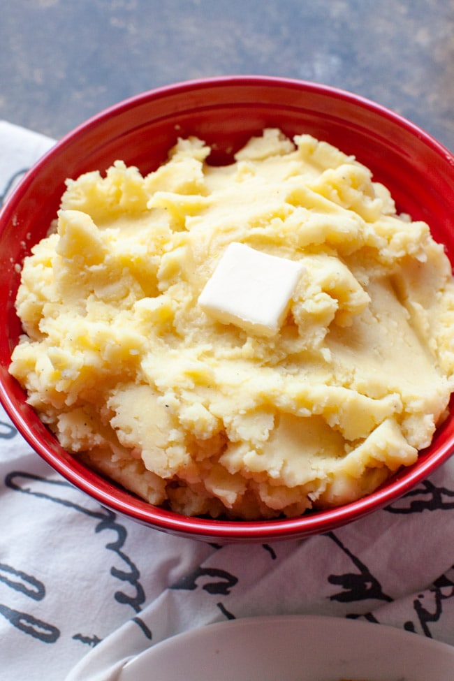How to Make the Perfect Mashed Potatoes A large red bowl with mashed potatoes and butter on top