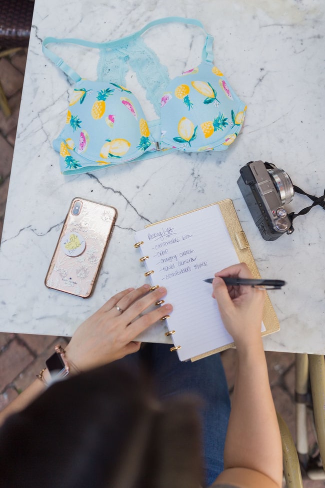 Taking notes for travel thelittlekitchen.net a woman writing notes on a notebook with a camera, phone and bra on a table