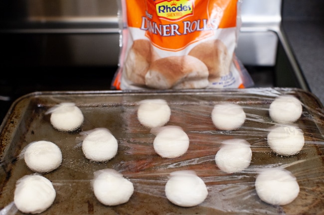 Stuffed Bagel Bites from thelittlekitchen.net frozen dinner rolls defrosting on a baking sheet covered in plastic with a bag of Rhodes Yeast Dinner Rolls in the background