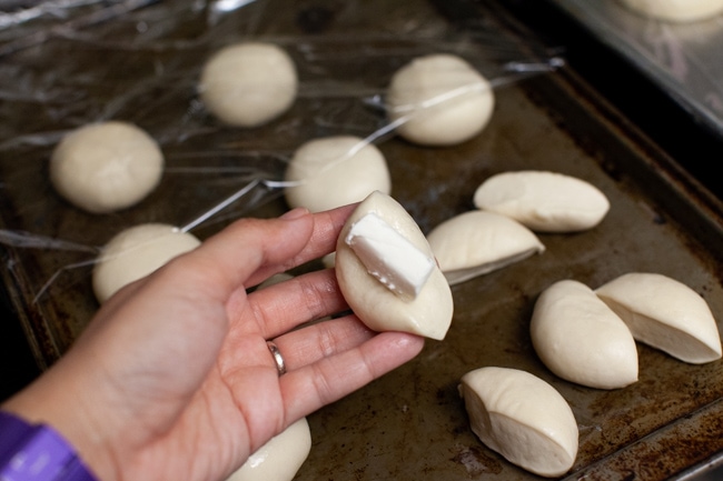 Stuffed Bagel Bites from thelittlekitchen.net one Rhodes dinner rolls cut in half with cream cheese on a hand with other rolls on a baking sheet in the background